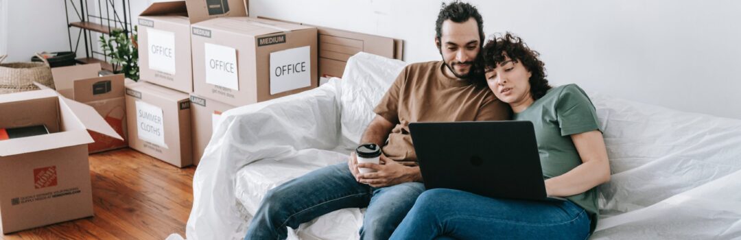 couple sitting on couch using laptop with moving boxes near by